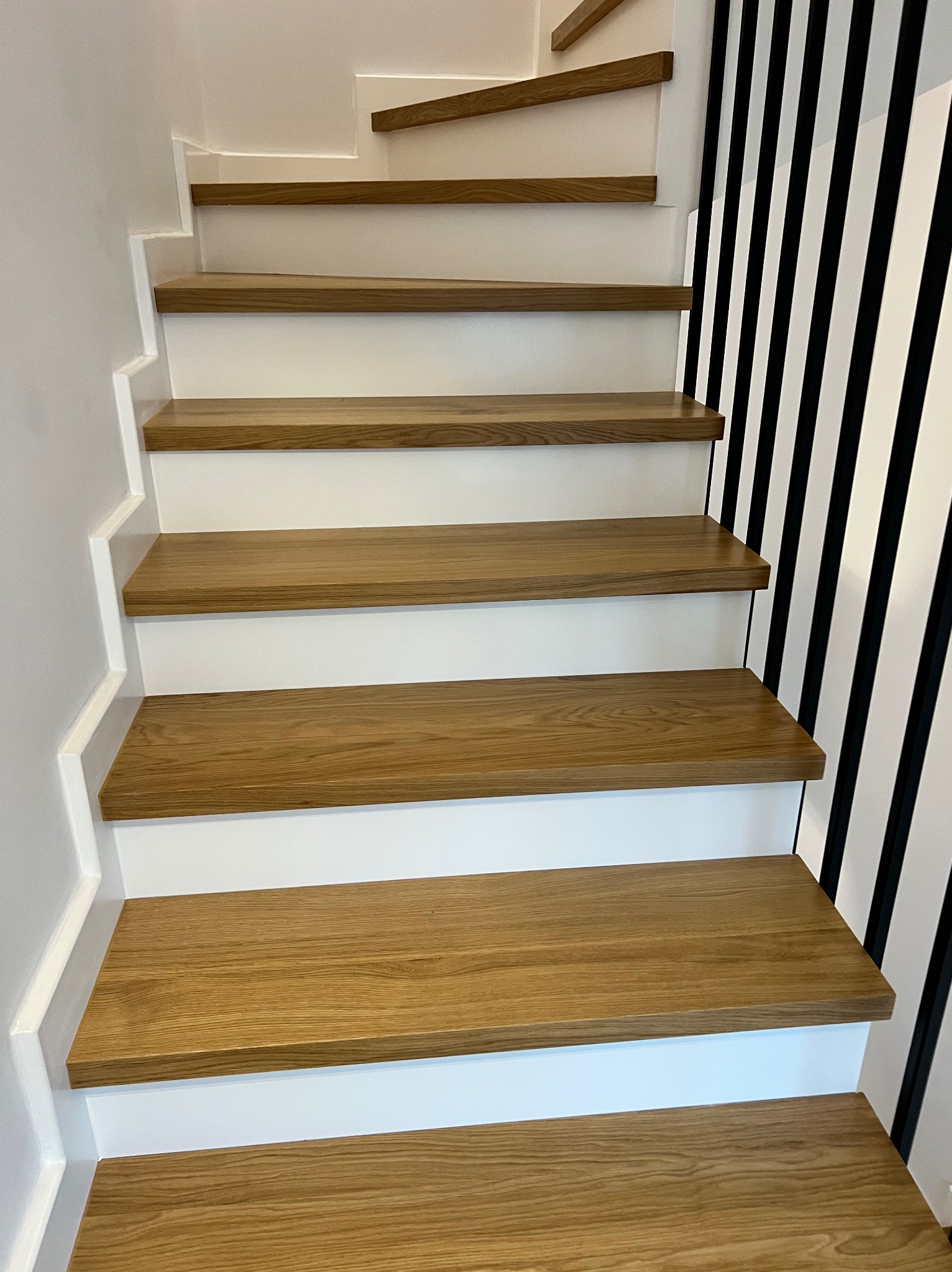 Nowoczesne schody z dębowymi stopniami i białymi podstopnicami, kontrastujące z czarno-białą, pionową dekoracją ściany. Minimalistyczny design.