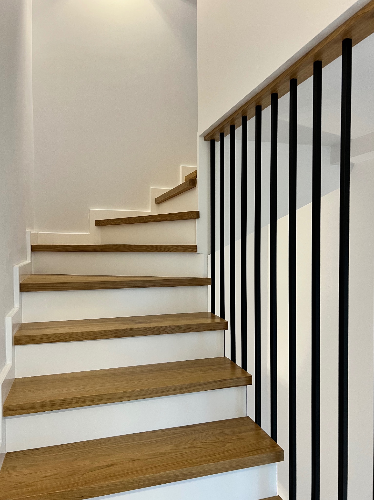 Nowoczesne schody drewniane z białymi podstopnicami i czarną, minimalistyczną balustradą. Jasne oświetlenie podkreśla naturalny odcień drewna.