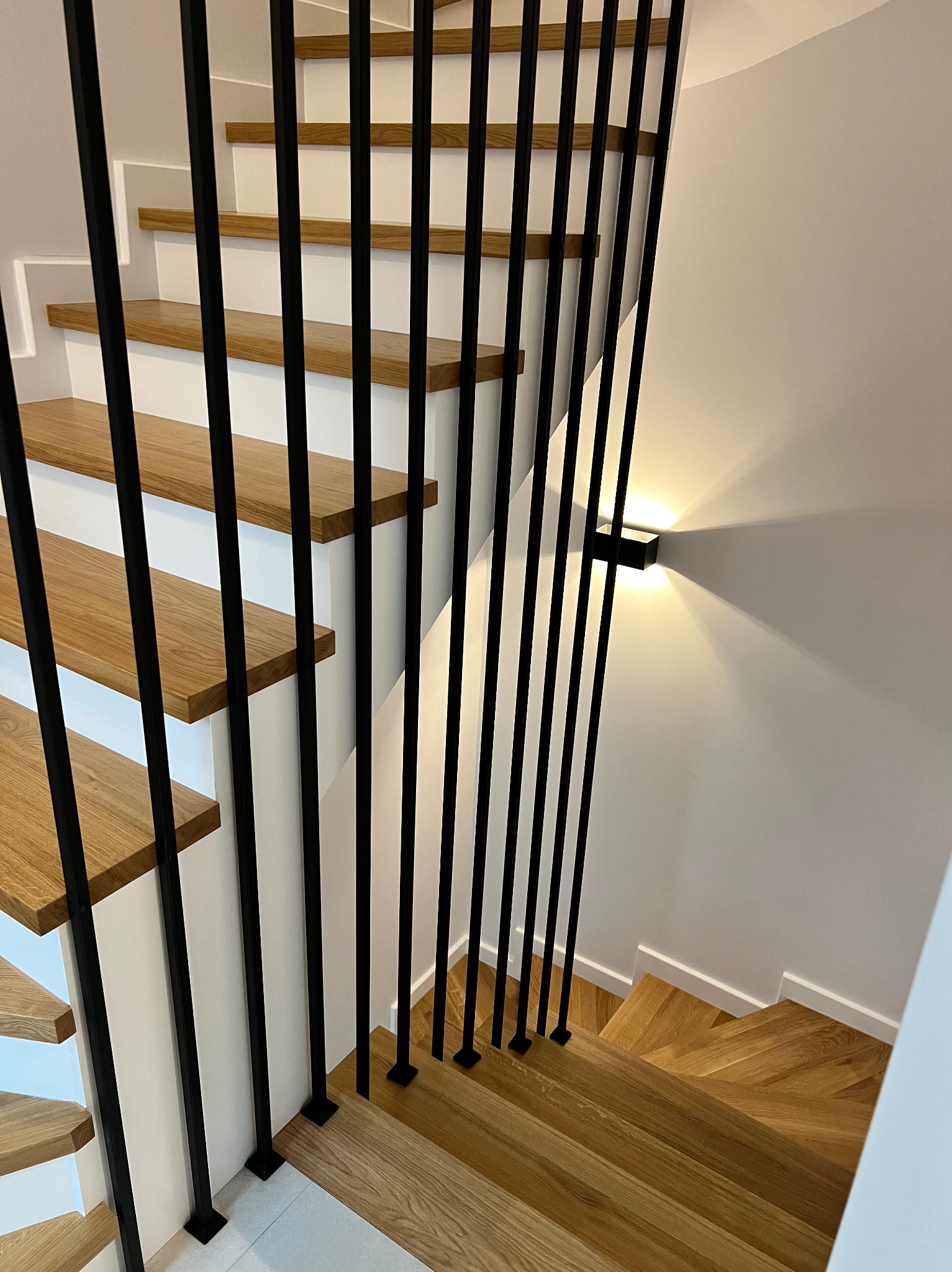 Nowoczesne schody z drewnianymi stopniami i czarnymi, pionowymi elementami w Rzeszotarach. Minimalistyczne oświetlenie ścienne podkreśla design.
