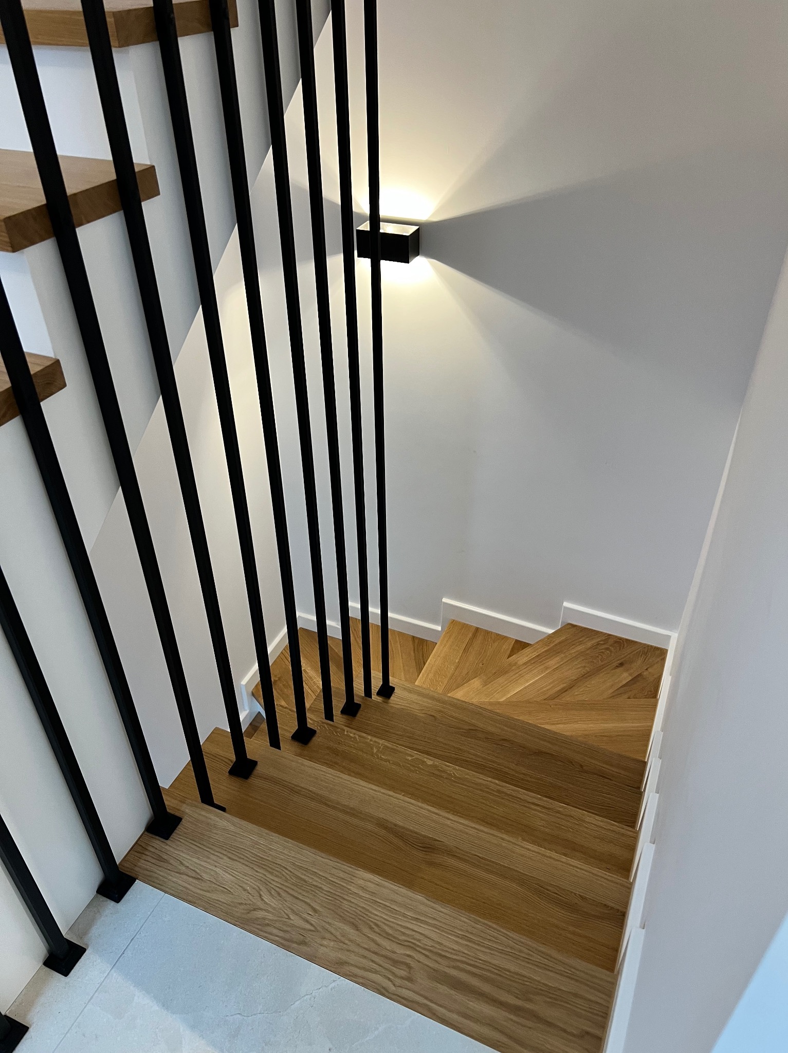Nowoczesne schody z drewnianymi stopniami i czarnymi, pionowymi balustradami, oświetlone kinkietem na białej ścianie. Widok z góry.