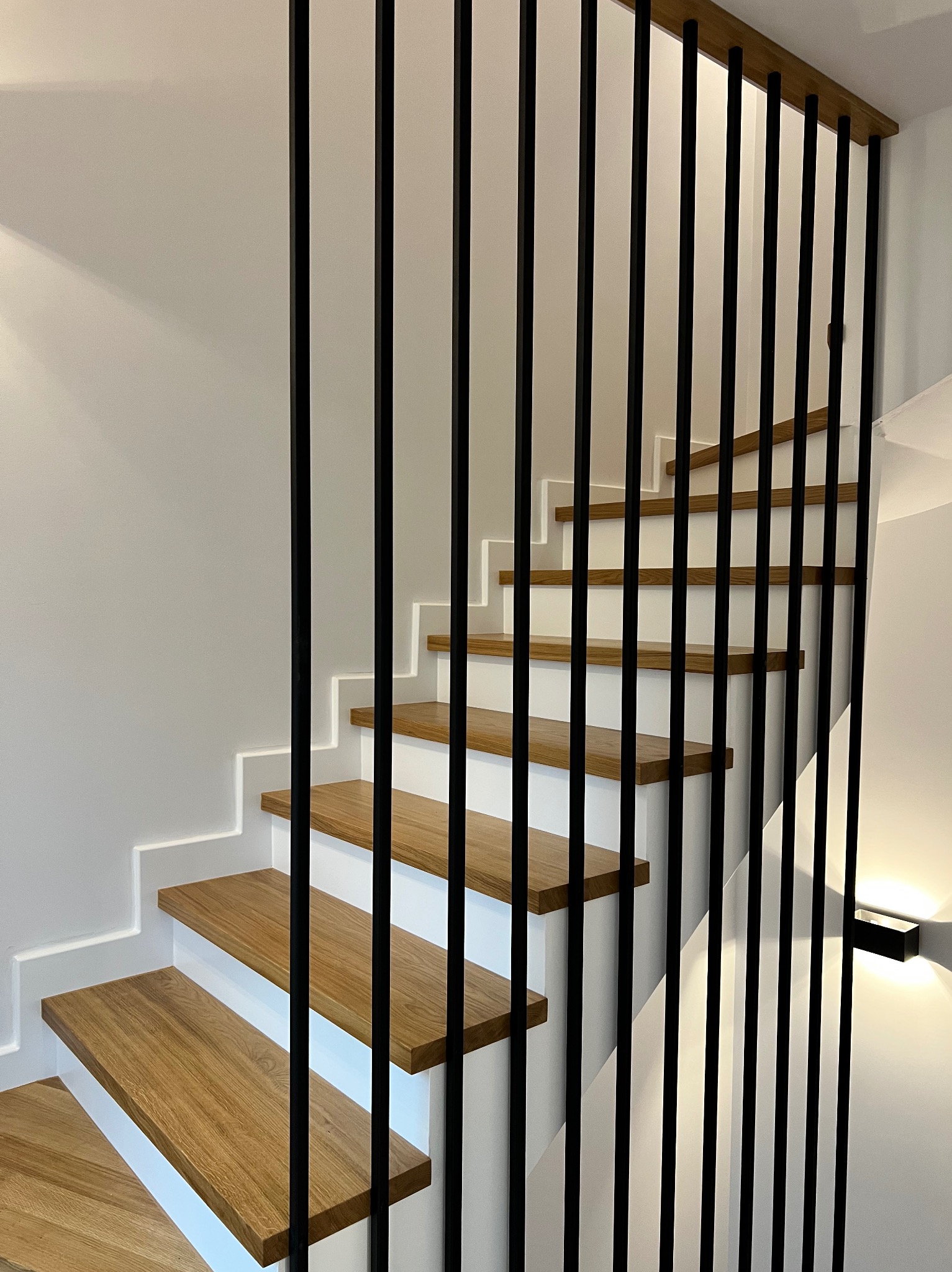 Nowoczesne schody z drewnianymi stopniami i minimalistyczną, czarną balustradą z pionowych prętów. Oświetlenie LED podkreśla białe podstopnice.