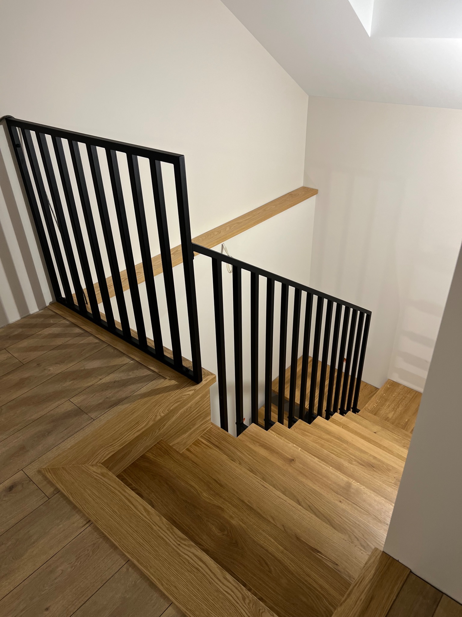 Nowoczesne schody z jasnego drewna z czarną, minimalistyczną balustradą. Widok z góry, podkreślający geometryczny wzór parkietu na podeście.