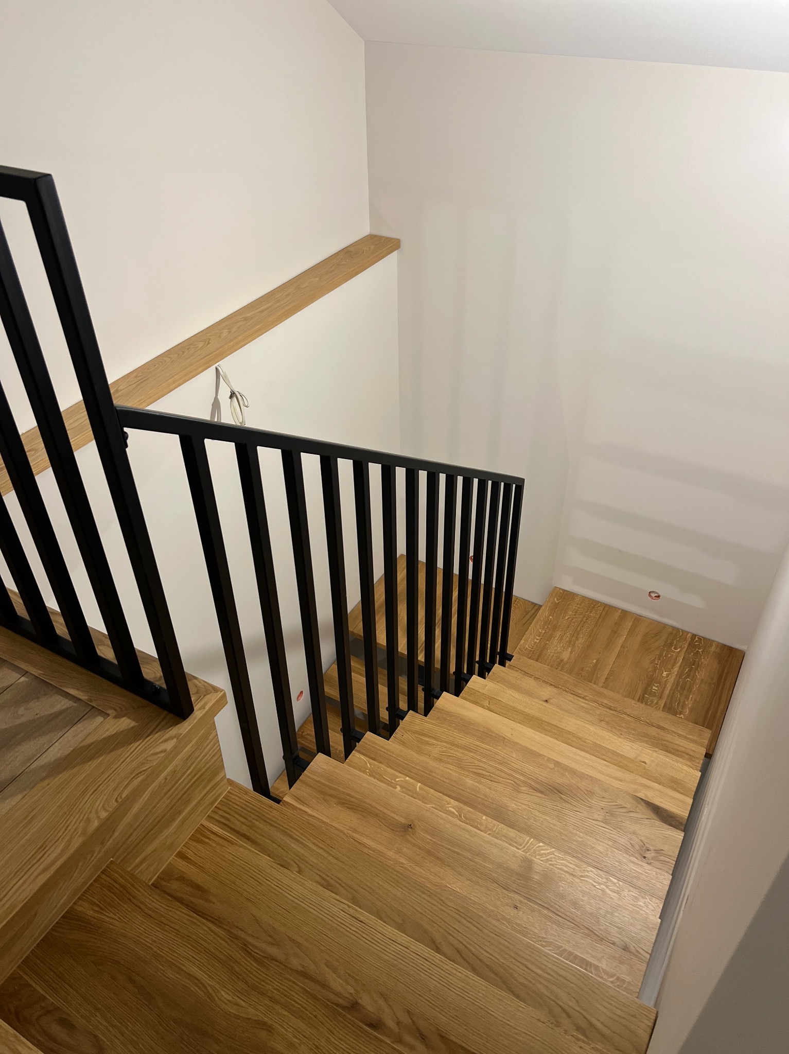 Nowoczesne schody z drewna z czarną, metalową balustradą, widok z góry. Jasne drewno stopni kontrastuje z ciemną balustradą i białymi ścianami.