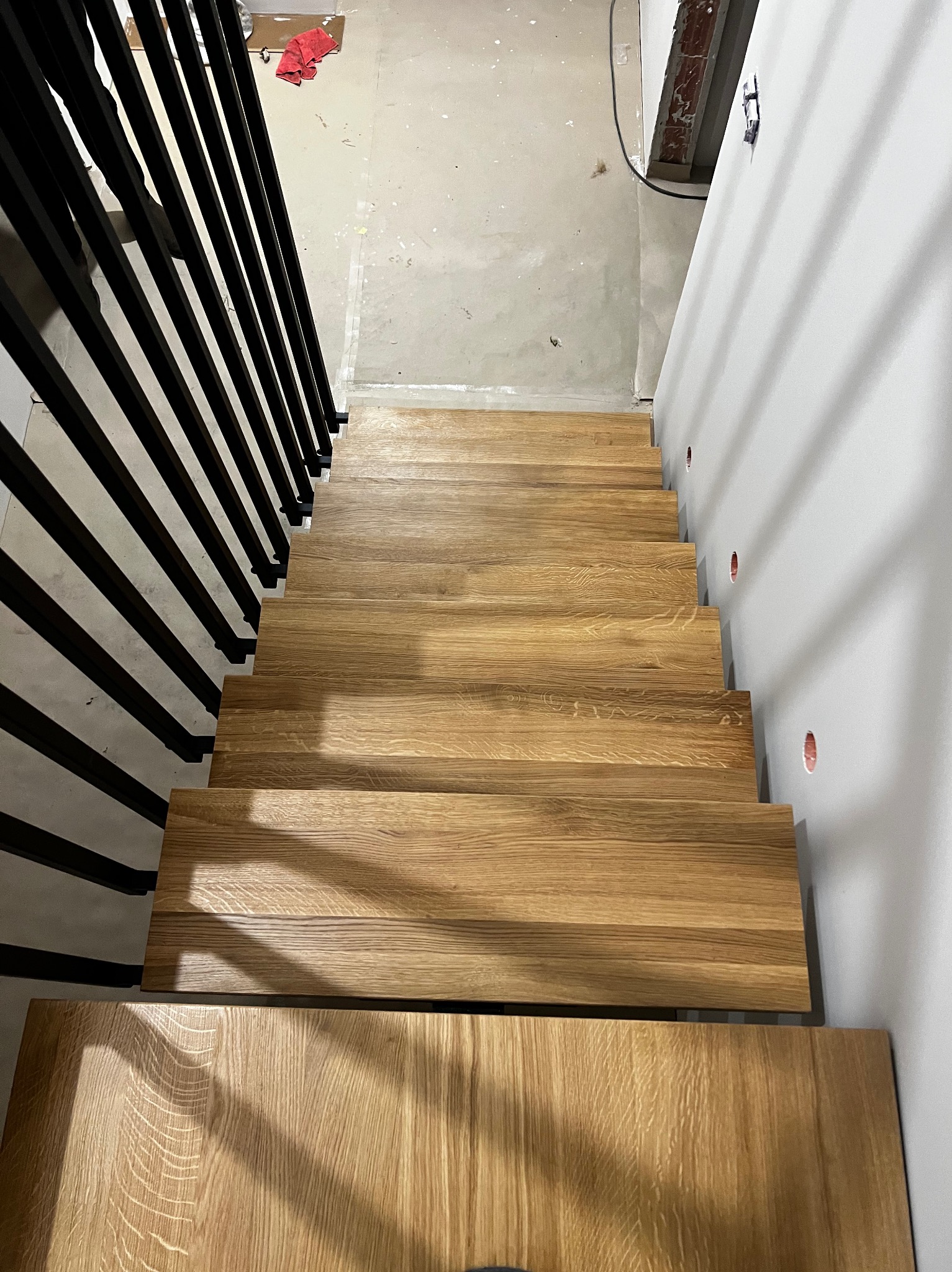 Nowoczesne schody z jasnego drewna, widok z góry, z czarną, minimalistyczną poręczą. Świeżo wykończone, z widocznymi śladami prac wykończeniowych na ścianach.