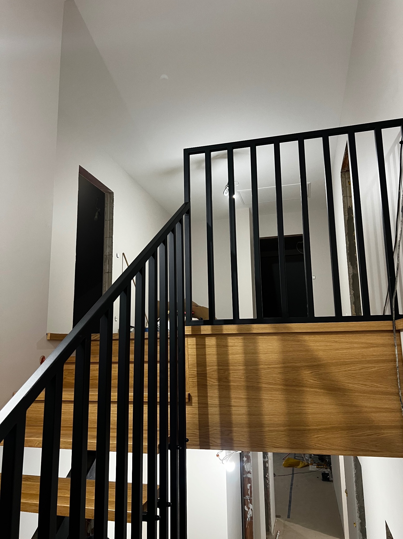 Nowoczesne schody z drewnianymi stopniami i czarną, metalową balustradą. Ujęcie z dołu, widoczne detale wykończenia i oświetlenie.