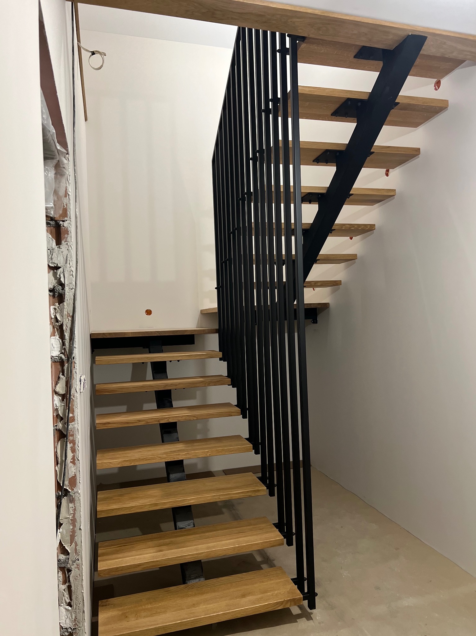 Nowoczesne schody z drewnianymi stopniami i czarną, minimalistyczną konstrukcją metalową. Widoczne surowe wykończenie ściany, kontrastujące z gładką powierzchnią.