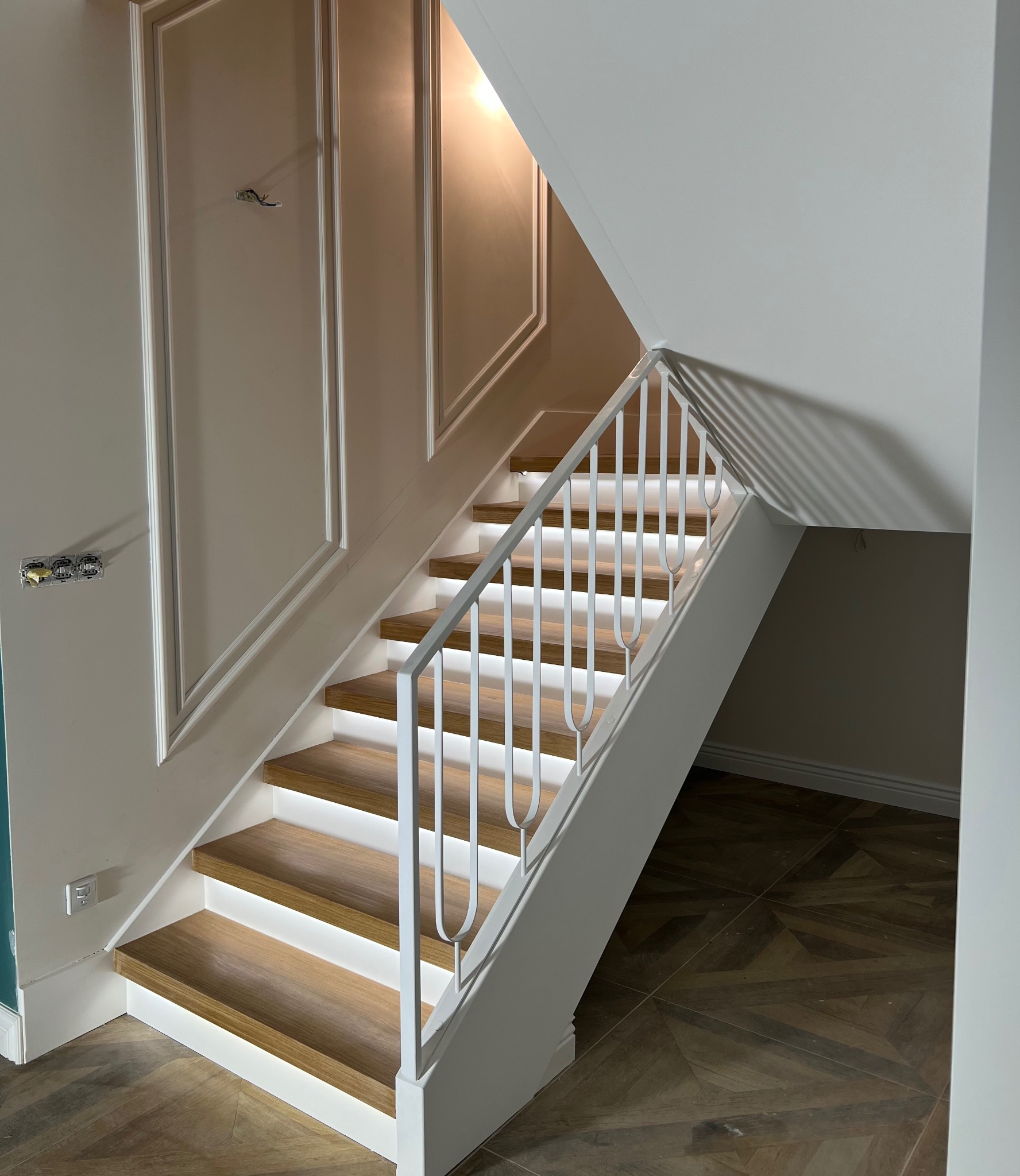 Nowoczesne schody z drewnianymi stopniami, podświetleniem LED i białą, metalową balustradą. Ściana z dekoracyjnymi listwami. Parkiet w jodełkę na podłodze.