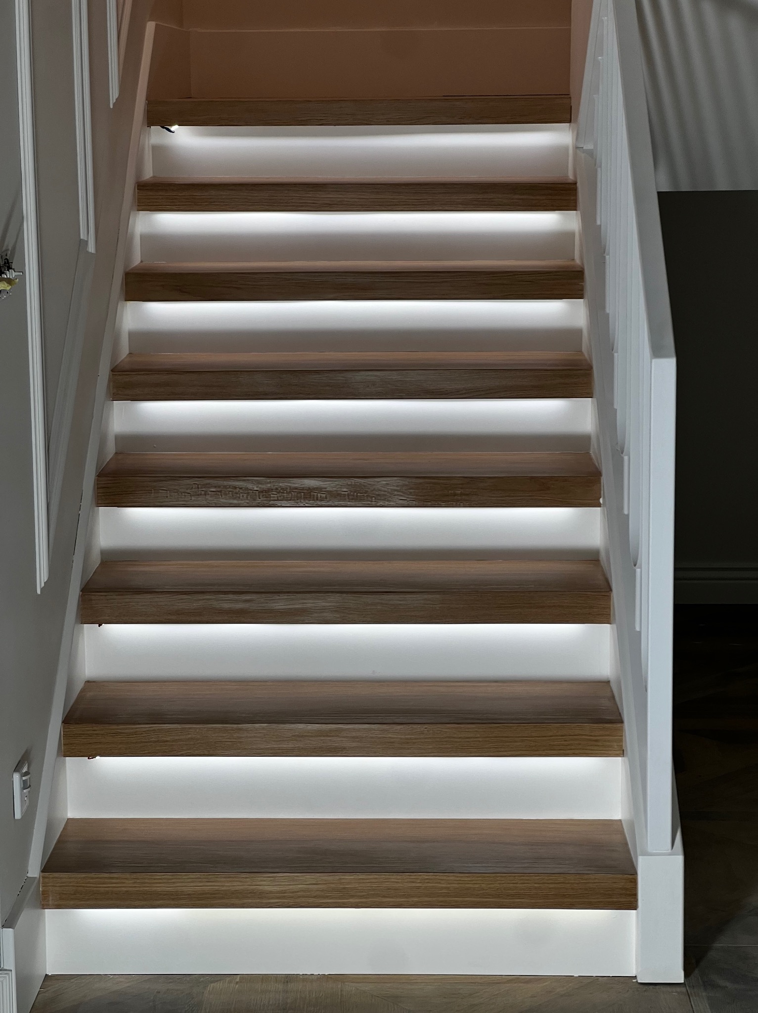Nowoczesne schody z drewna z białymi podstopnicami i podświetleniem LED, widok z dołu. Minimalistyczny design, jasne oświetlenie, eleganckie wykończenie.