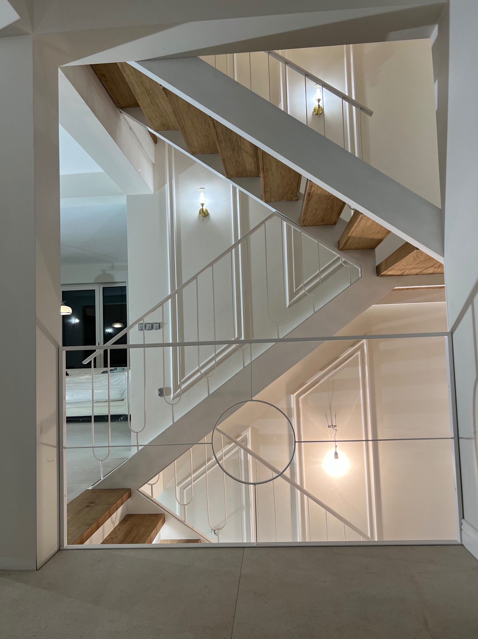 Białe schody z drewnianymi stopniami, widoczne przez kwadratowe wejście. Minimalistyczny design z oświetleniem ściennym i metalową balustradą.