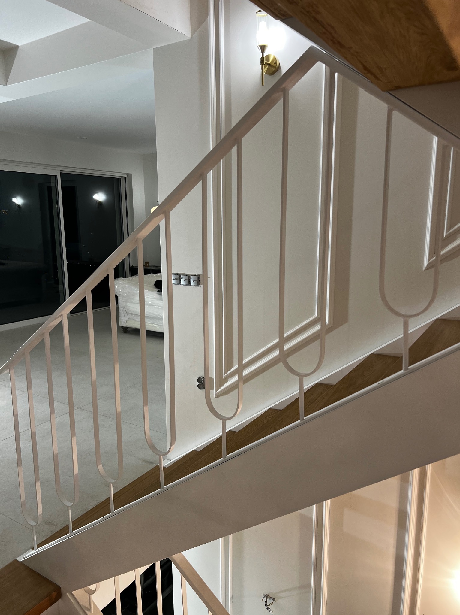 Nowoczesne, białe poręcze schodowe z dekoracyjnymi elementami w kształcie litery U, widoczne z góry, z drewnianymi stopniami i jasnym wnętrzem w tle.