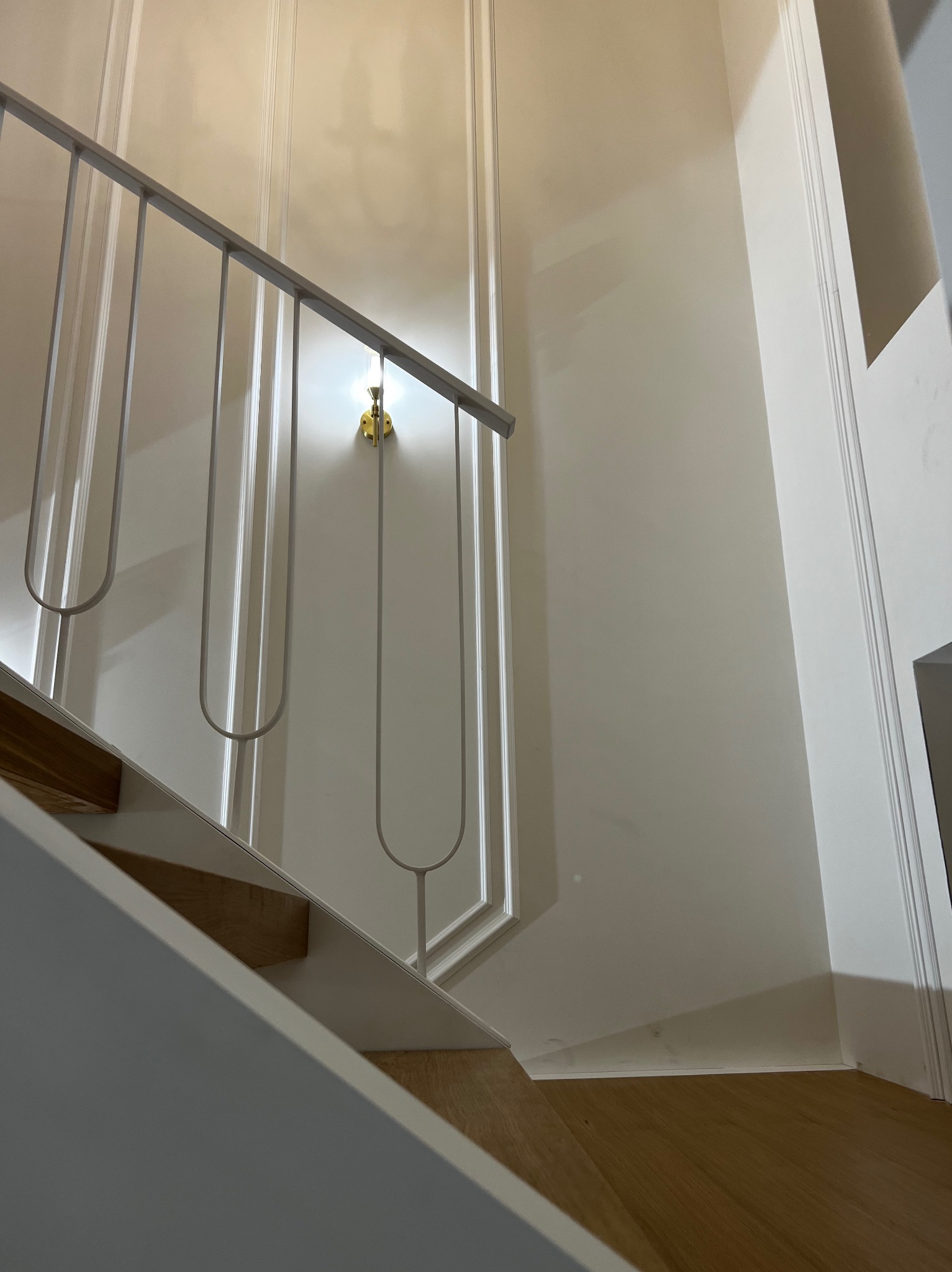 Nowoczesne schody z drewnianymi stopniami i minimalistyczną, metalową poręczą na tle białej ściany z dekoracyjnymi listwami i złotym kinkietem.