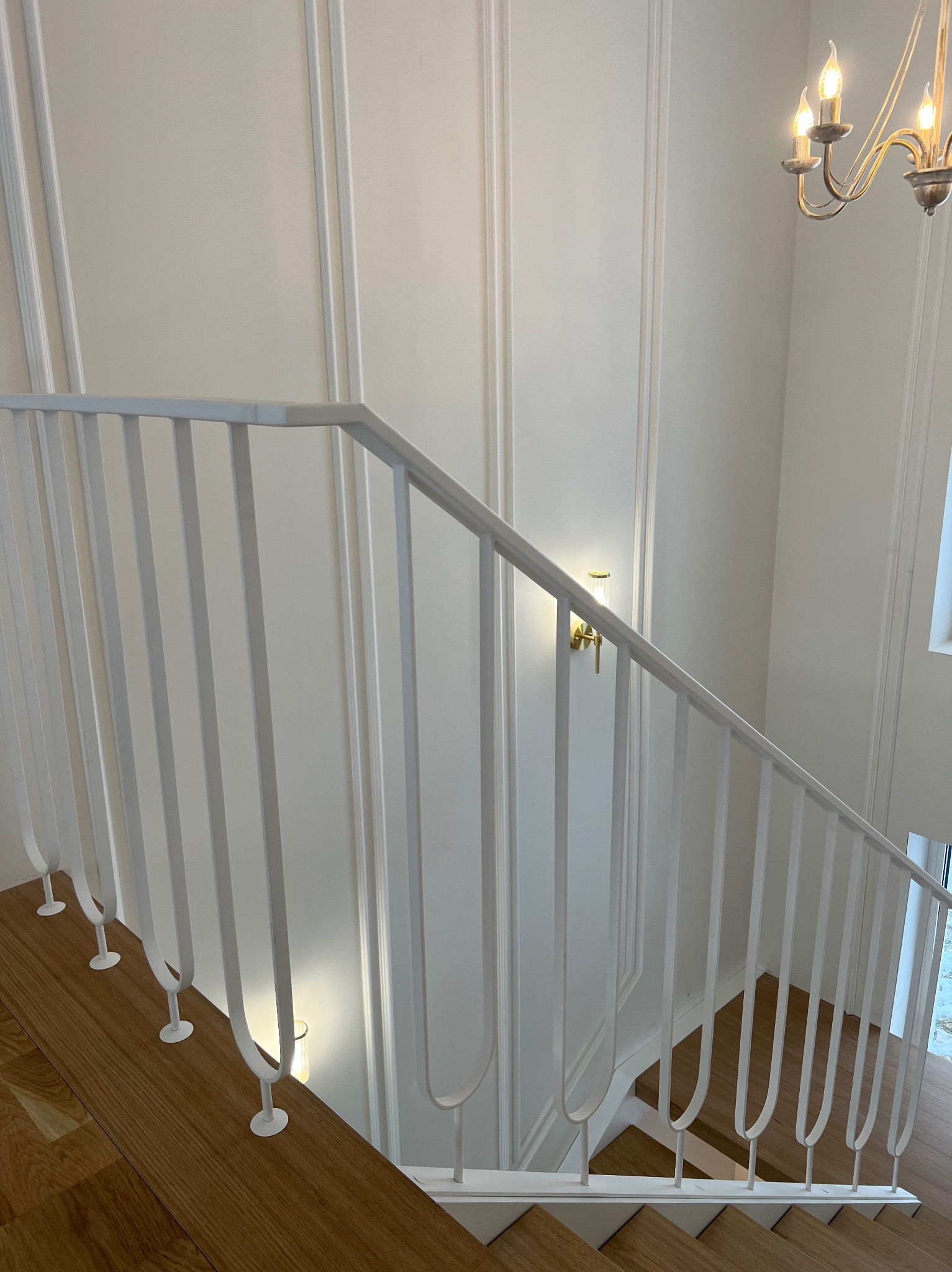 Nowoczesne schody z drewnianymi stopniami i białą, metalową balustradą o minimalistycznym designie. Ściany zdobione sztukaterią, oświetlenie punktowe i żyrandol.