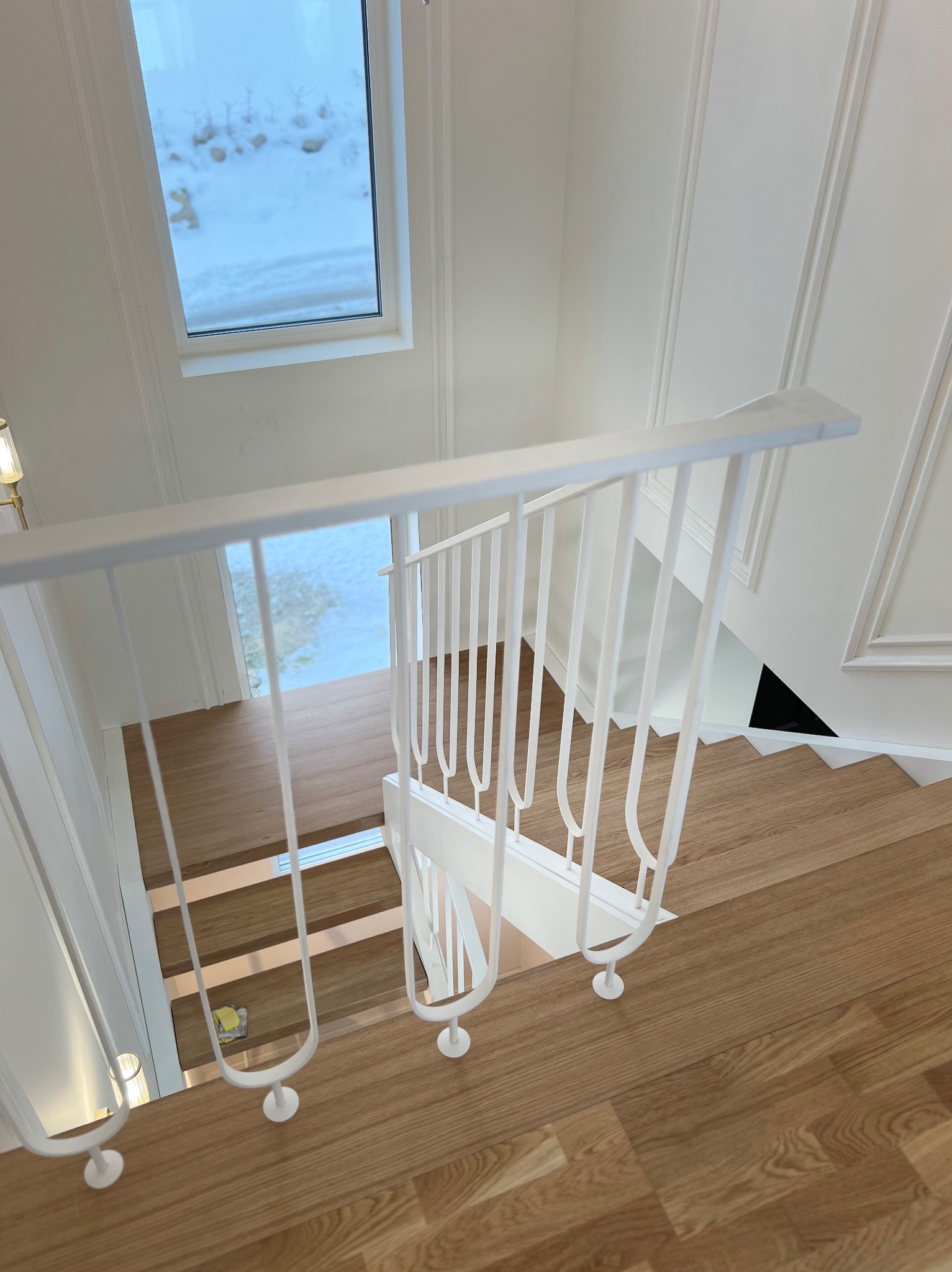 Jasne, drewniane schody z białymi, metalowymi poręczami, widziane z góry. W tle okno z widokiem na zimowy krajobraz. Minimalistyczny design.