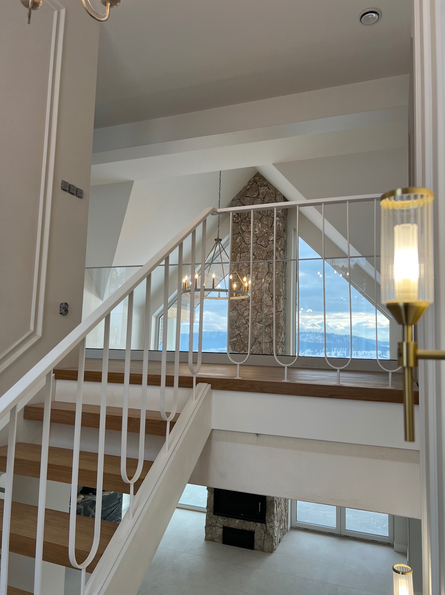 Jasne, nowoczesne wnętrze z drewnianymi schodami i białymi poręczami. Widok na salon z kamiennym kominkiem i dużym oknem z widokiem na góry. Elegancki żyrandol.