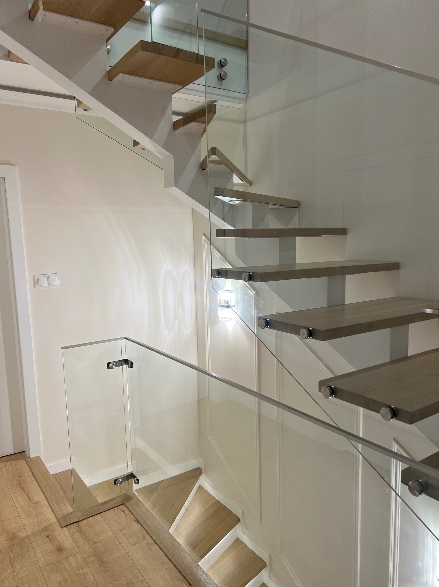 Nowoczesne schody z drewnianymi stopniami i szklaną balustradą, mocowaną punktowo. Minimalistyczny design, jasne wnętrze, widok z dołu.