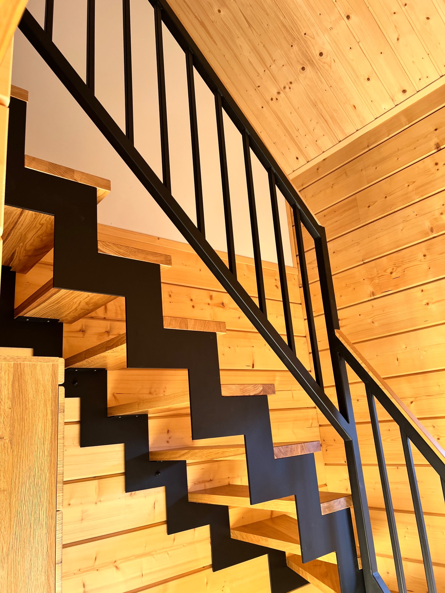 Nowoczesne schody z drewnianymi stopniami i czarną, metalową konstrukcją, w jasnym wnętrzu wykończonym drewnianą boazerią. Ujęcie z dołu, podkreślające geometrię.