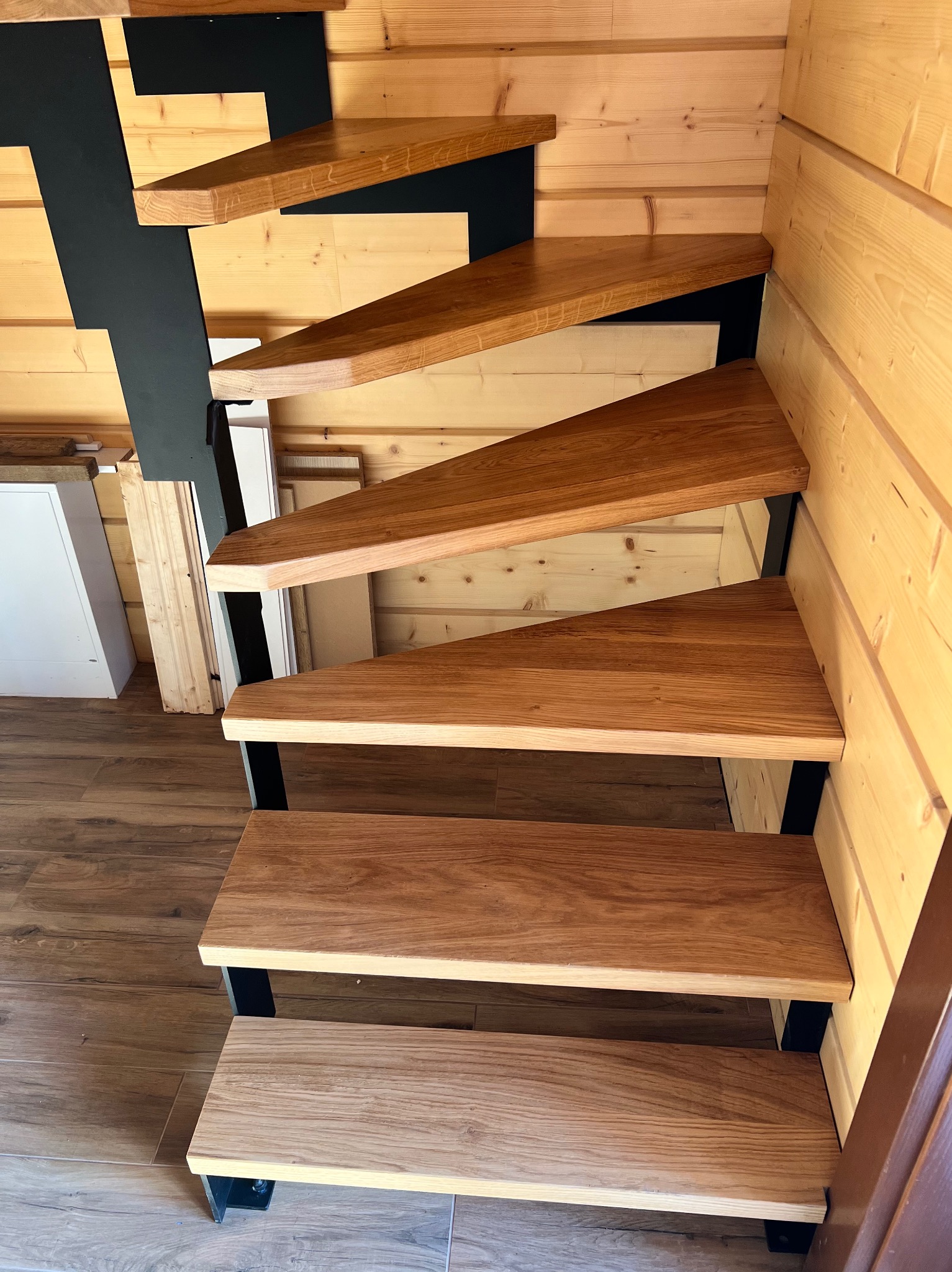 Nowoczesne schody z drewnianymi stopniami i czarną, metalową konstrukcją w domu z boazerią. Widok z dołu, ukazujący detale wykończenia i połączenie materiałów.