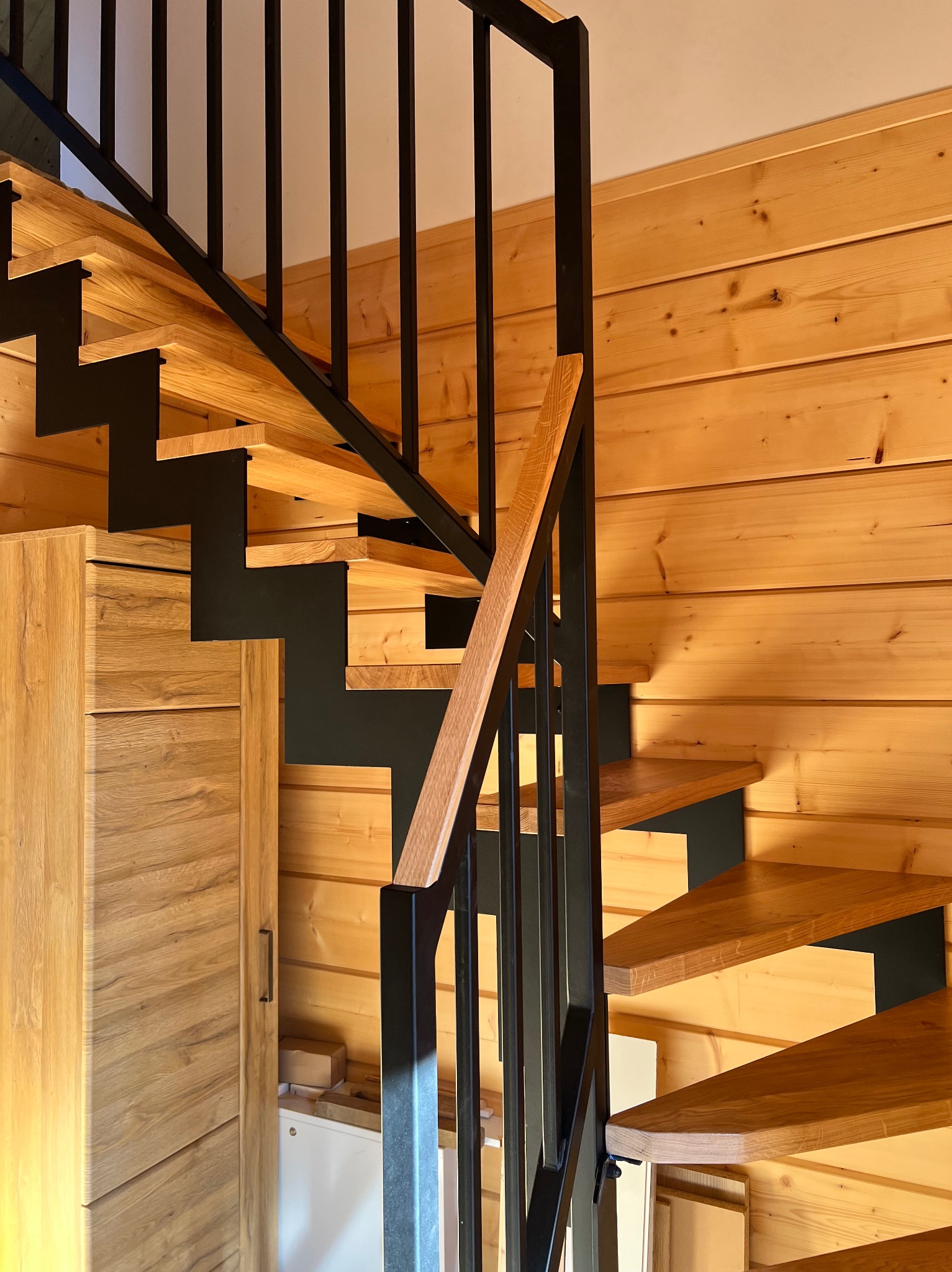 Nowoczesne schody z drewnianymi stopniami i czarną, metalową konstrukcją, wkomponowane w drewniane wnętrze domu. Widoczna szafka pod schodami.