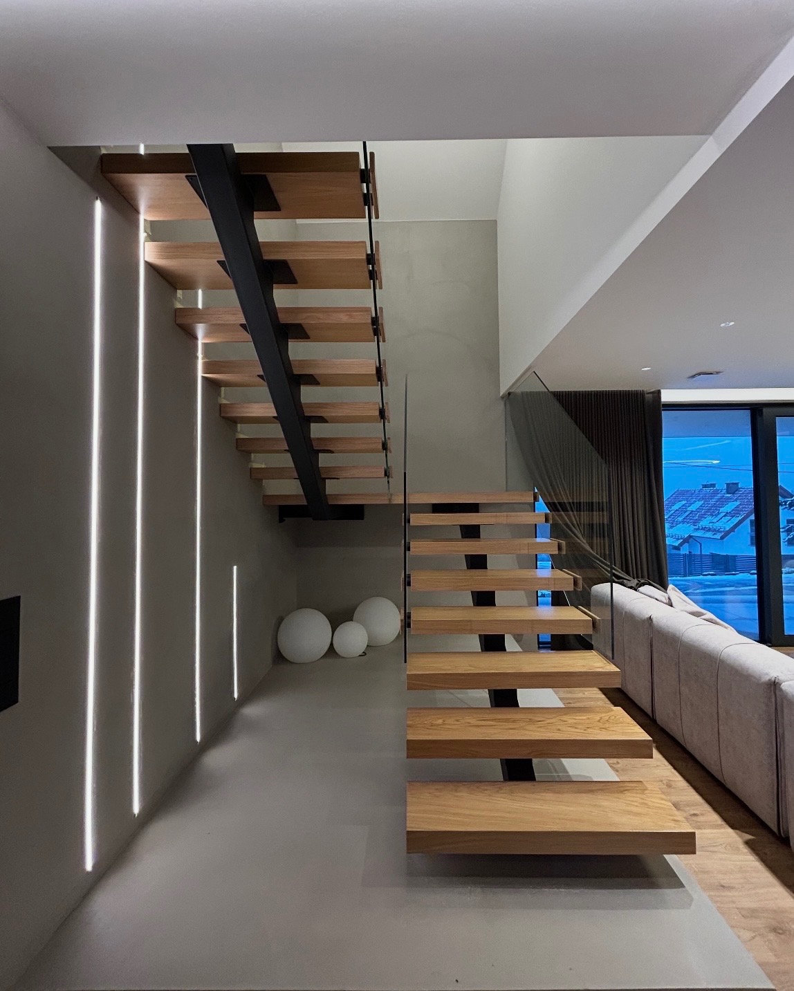 Nowoczesne schody drewniane z czarnymi wspornikami i szklaną balustradą, oświetlone pionowymi liniami LED na ścianie. Minimalistyczny design.