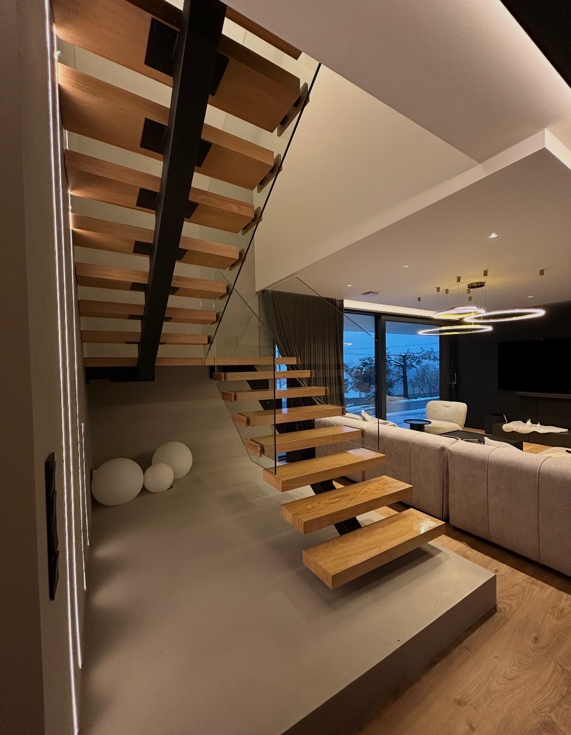 Nowoczesne schody drewniane z czarną konstrukcją i szklanymi panelami, w jasnym wnętrzu z widokiem na zewnątrz. Minimalistyczny design, ciepłe oświetlenie.