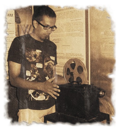 Mężczyzna w okularach, w koszulce z nadrukiem komiksowym, obsługuje stary projektor filmowy z taśmą, na tle ściany z plakatami.