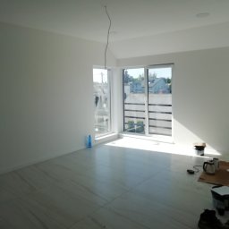 Fel-Bud Przemysław Feliński - Wnętrze świeżo pomalowanego pokoju z białymi ścianami i jasną podłogą z płytek, widoczne narzędzia malarskie i butelka wody, kabel zwisający z sufitu.