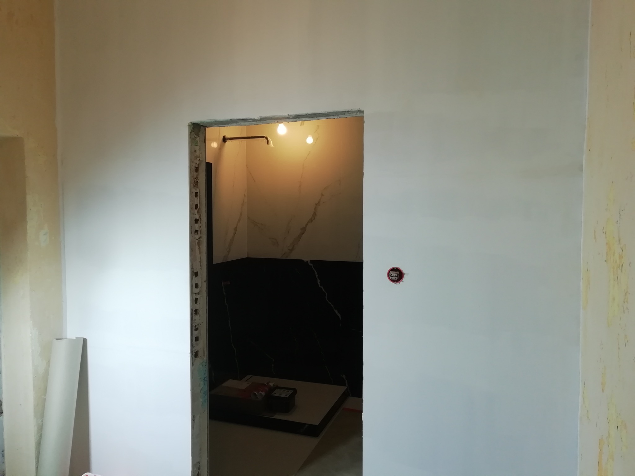 Świeżo pomalowana ściana w jasnym odcieniu z widocznym otworem drzwiowym prowadzącym do wykończonej łazienki z czarnymi płytkami imitującymi marmur i białymi płytkami na ścianie, w tle widoczne...