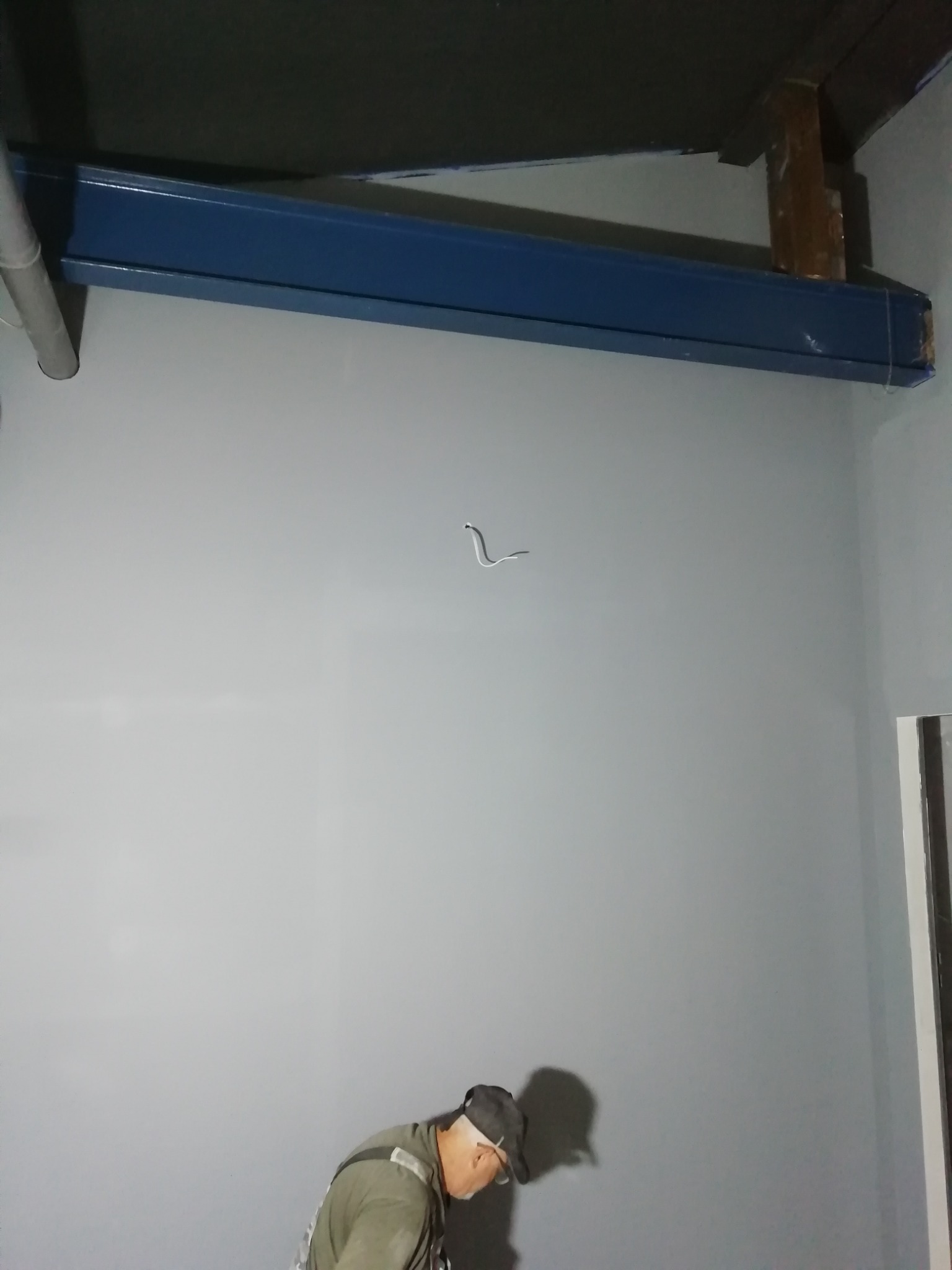 Malowanie szarej ściany w pomieszczeniu z niebieską belką konstrukcyjną pod sufitem; widoczny fragment instalacji elektrycznej i pracownik w dolnej części kadru.