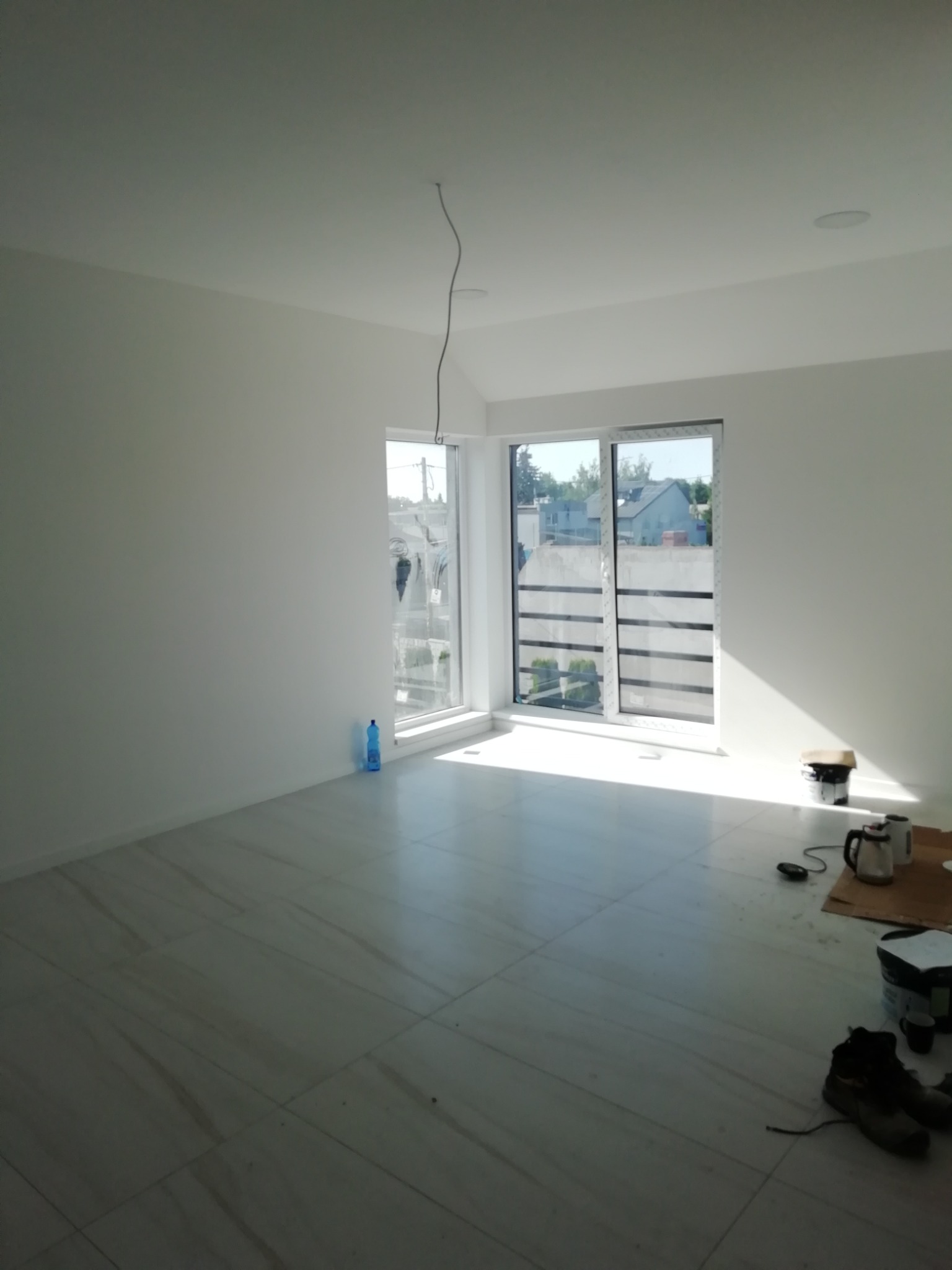 Wnętrze świeżo pomalowanego pokoju z białymi ścianami i jasną podłogą z płytek, widoczne narzędzia malarskie i butelka wody, kabel zwisający z sufitu.