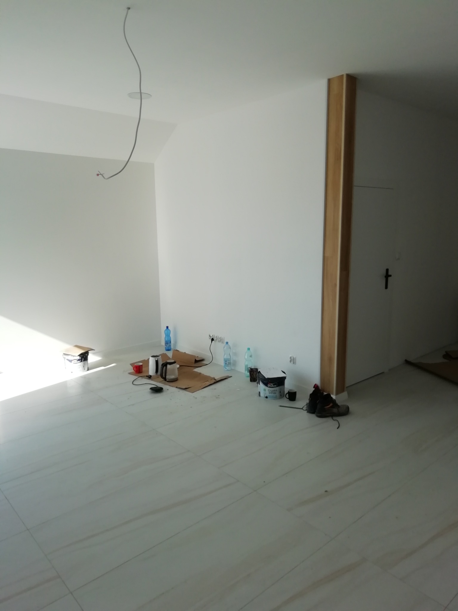 Pokój w trakcie malowania, widoczne białe ściany, podłoga wyłożona jasnymi płytkami imitującymi marmur, puszka farby Dulux Crystal White, buty robocze, kabel zwisający z sufitu, zakryte kartonami...