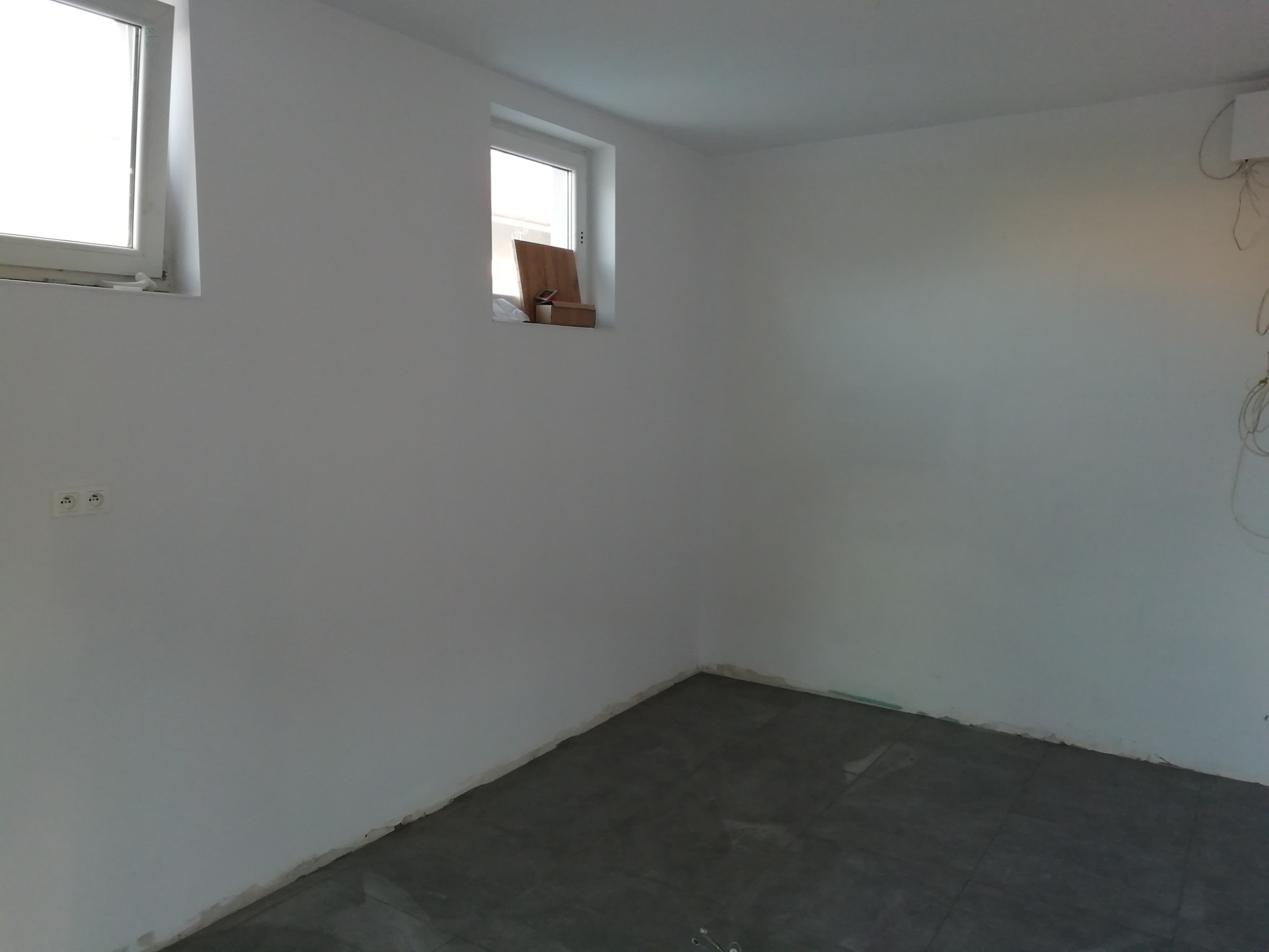 Pomalowane na biało wnętrze pokoju z szarymi płytkami podłogowymi, widoczne dwa okna i gniazdka elektryczne.