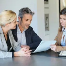 Para w średnim wieku, kobieta w jasnym żakiecie obejmująca mężczyznę, konsultuje dokumenty z doradcą finansowym przy stole w minimalistycznym wnętrzu.