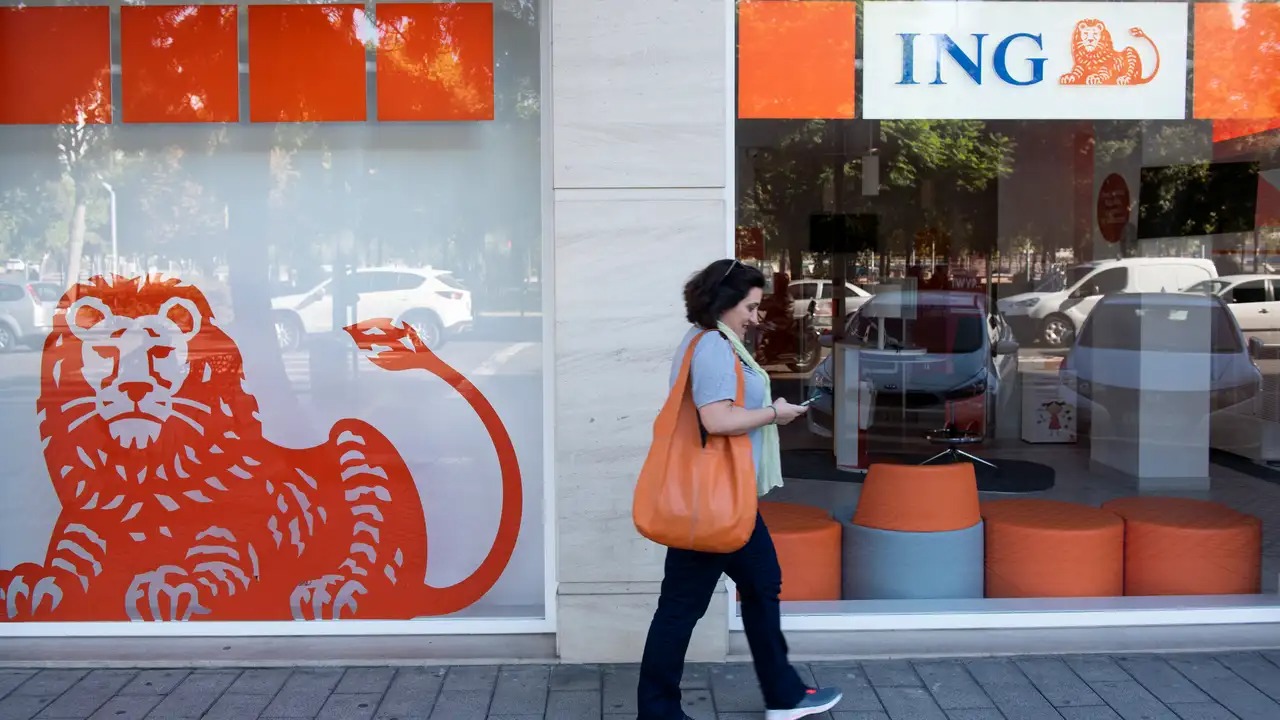 Kobieta z pomarańczową torbą przechodzi obok witryny banku z logo lwa w kolorze pomarańczowym, przeglądając telefon. W tle widoczne zaparkowane samochody.