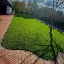 Starannie utrzymany trawnik graniczący z czerwoną kostką brukową ułożoną w jodełkę, cień drzewa na trawie, dom z białymi ścianami w tle, błękitne niebo z chmurami.