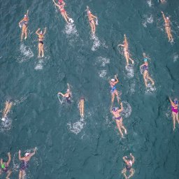 Aqua Champion - Widok z góry na grupę osób pływających w otwartym morzu w kolorowych strojach i czepkach, z widocznymi falami i pianą tworzoną przez ruchy pływaków.