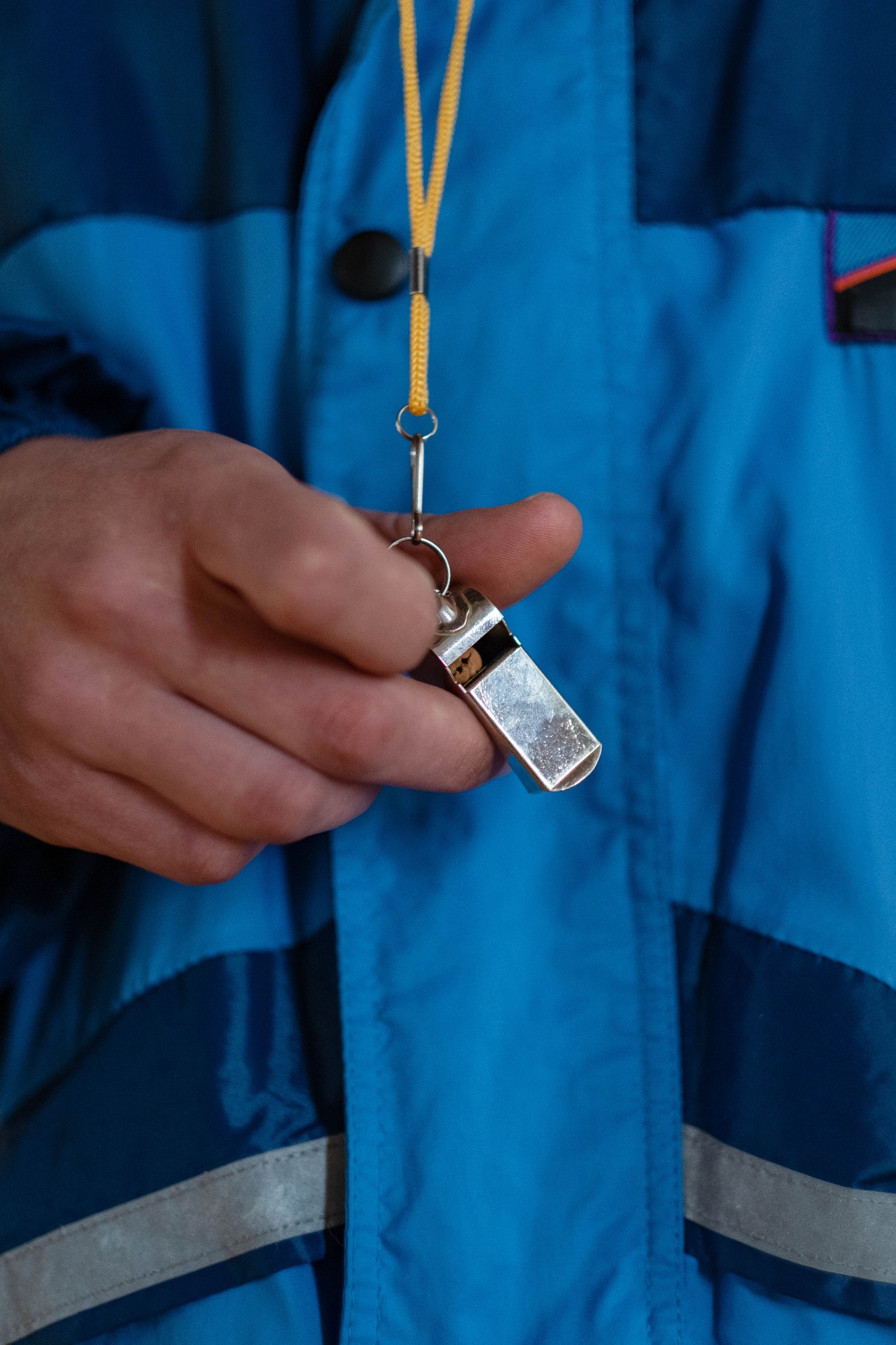 Dłoń trzymająca srebrny gwizdek na żółtym sznurku, na tle niebieskiej kurtki sportowej z odblaskowym paskiem.