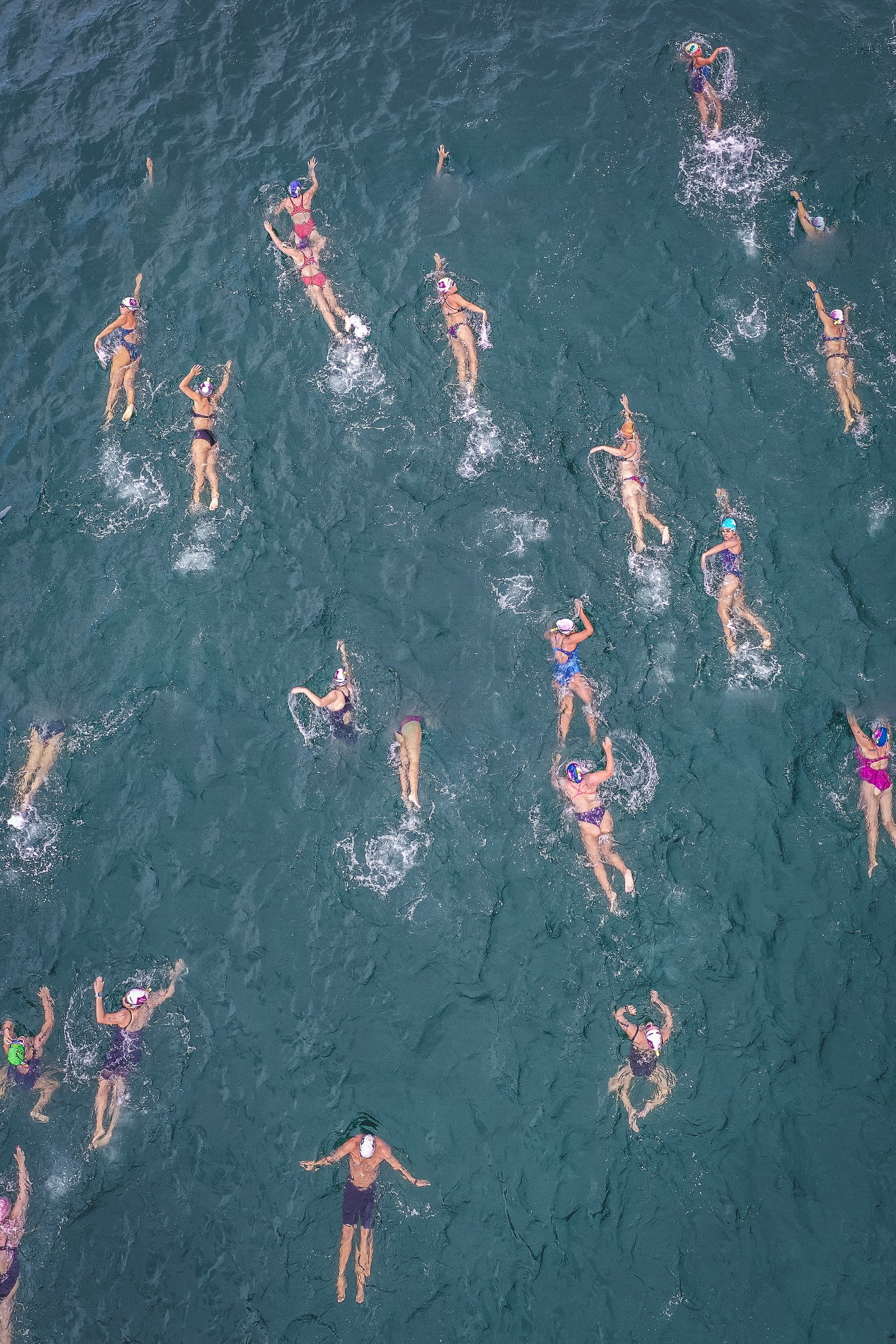 Widok z góry na grupę osób pływających w otwartym morzu w kolorowych strojach i czepkach, z widocznymi falami i pianą tworzoną przez ruchy pływaków.