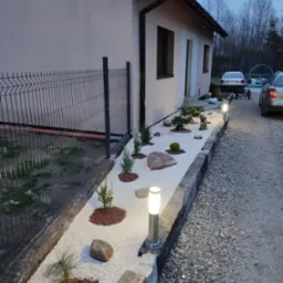 Ogród przy domu z białym żwirem, kamieniami, niskimi roślinami iglastymi i oświetleniem punktowym, widok na elewację budynku i ogrodzenie.