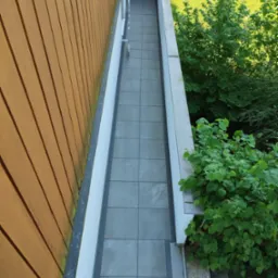 Widok z g&oacute;ry na wyłożony szarymi płytkami balkon z drewnianą ścianą po lewej stronie i zielenią po prawej, z widoczną konstrukcją balkonu powyżej i fragmentem trawnika w tle.