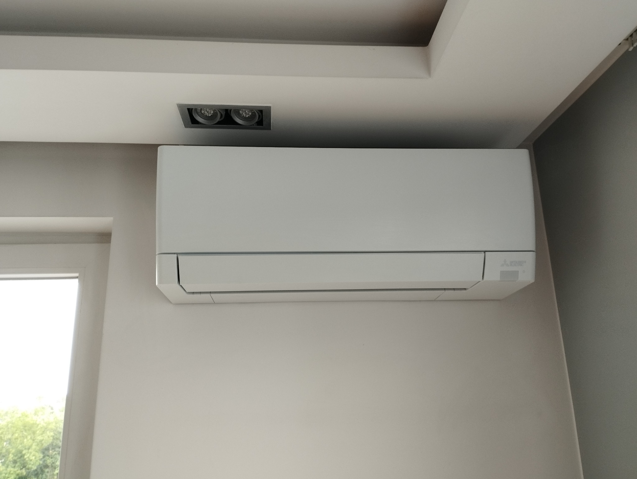 Biała jednostka klimatyzacji Mitsubishi Electric zamontowana na ścianie w jasnym pomieszczeniu z oknem, widoczne oświetlenie sufitowe.
