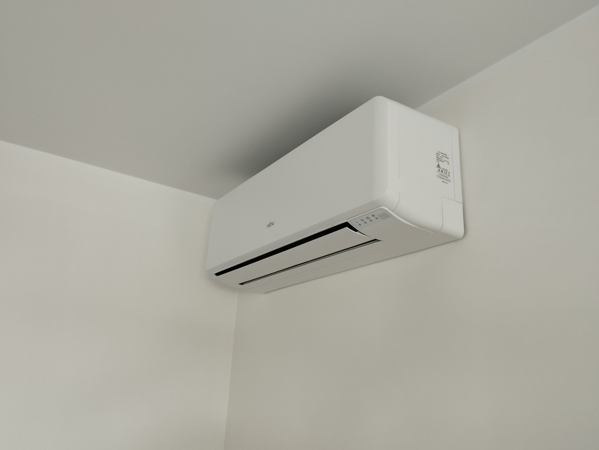Biała jednostka klimatyzacji Fujitsu zamontowana w rogu pomieszczenia z białymi ścianami i sufitem, widoczne detale panelu sterowania.