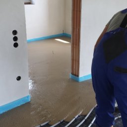 Posadzki anhydrytowe - Wylewanie płynnego jastrychu na ogrzewanie podłogowe w pomieszczeniu z widoczną izolacją przy ścianach oraz fragmentem pracownika w kombinezonie.