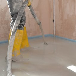 Posadzki anhydrytowe - Aplikacja wylewki samopoziomującej w pomieszczeniu z zabezpieczonymi ścianami, widoczne żółte gumowce pracownika i wąż doprowadzający masę.