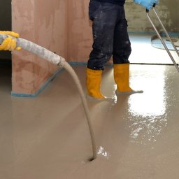 Posadzki anhydrytowe - Wylewanie beżowej masy na podłogę w pomieszczeniu, widoczne żółte kalosze i wąż doprowadzający mieszankę oraz narzędzie do rozprowadzania masy.