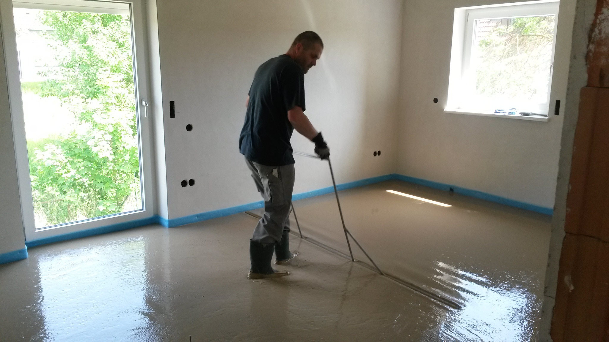 Aplikacja samopoziomującej wylewki w jasnym pomieszczeniu, widoczne narzędzie do rozprowadzania masy, niebieska taśma zabezpieczająca ściany, pracownik w gumowych butach i rękawicach.