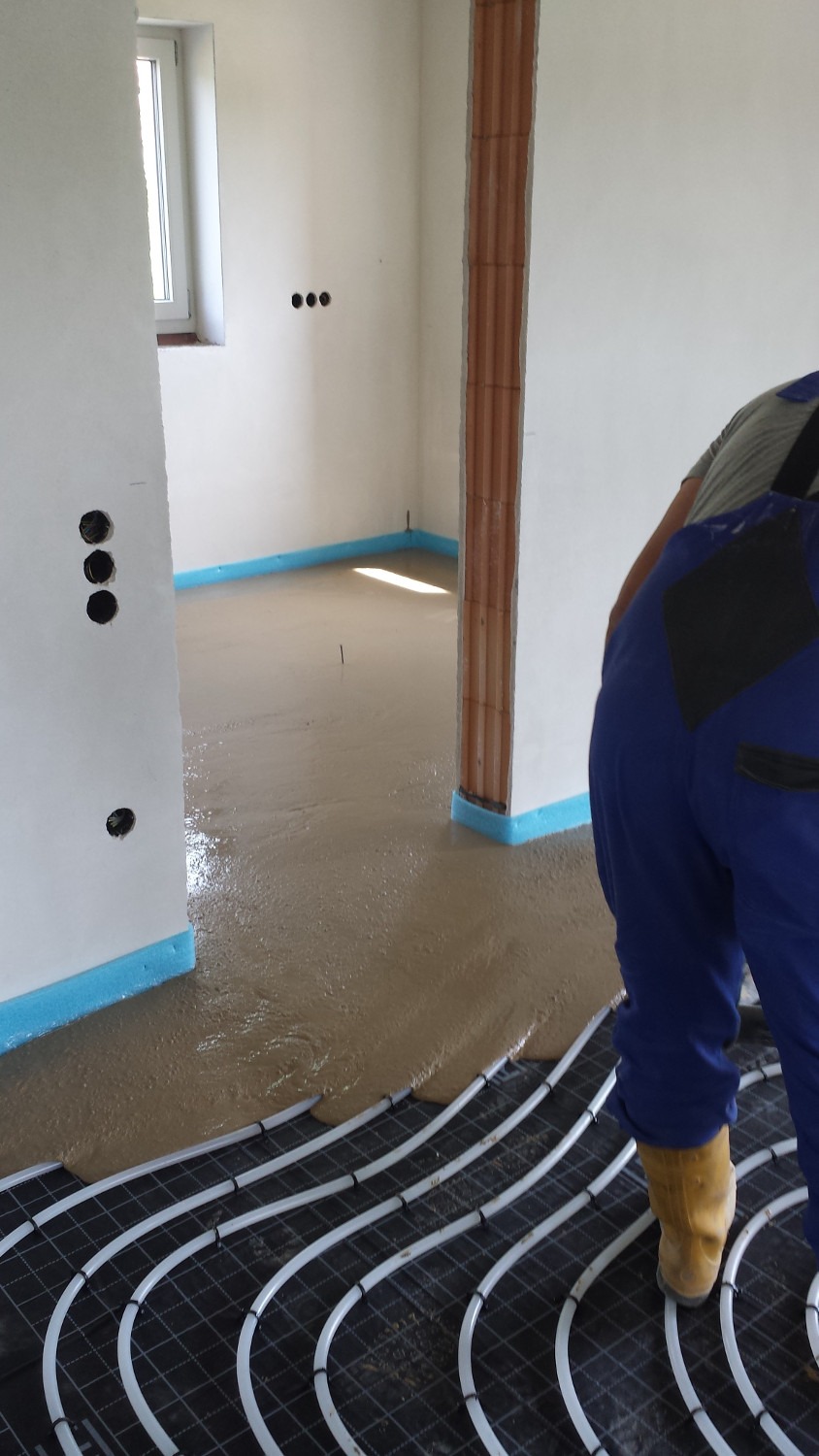 Wylewanie płynnego jastrychu na ogrzewanie podłogowe w pomieszczeniu z widoczną izolacją przy ścianach oraz fragmentem pracownika w kombinezonie.