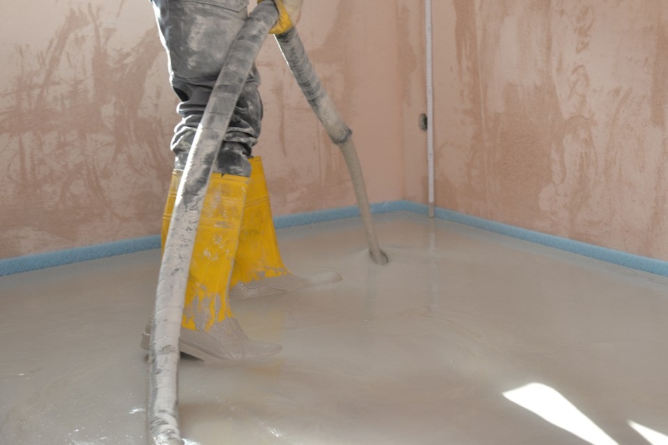 Aplikacja wylewki samopoziomującej w pomieszczeniu z zabezpieczonymi ścianami, widoczne żółte gumowce pracownika i wąż doprowadzający masę.