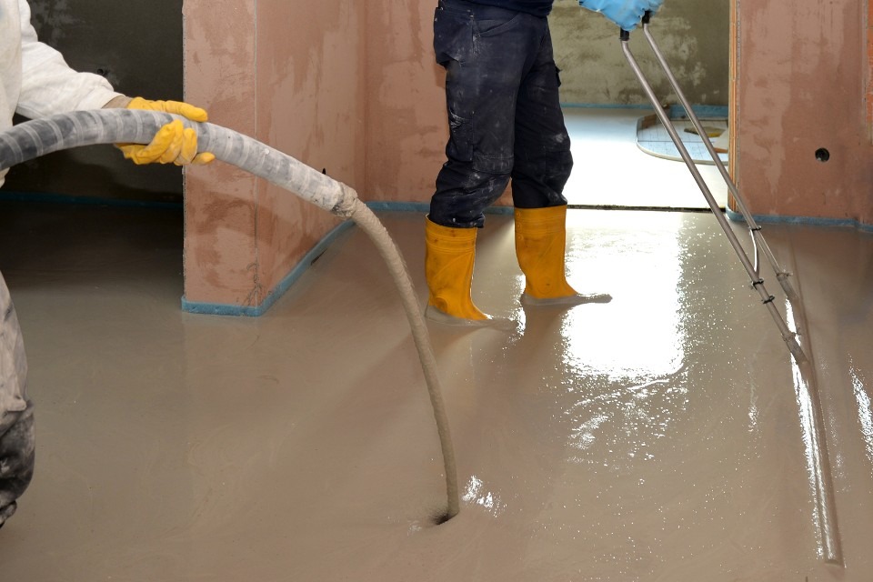 Wylewanie beżowej masy na podłogę w pomieszczeniu, widoczne żółte kalosze i wąż doprowadzający mieszankę oraz narzędzie do rozprowadzania masy.