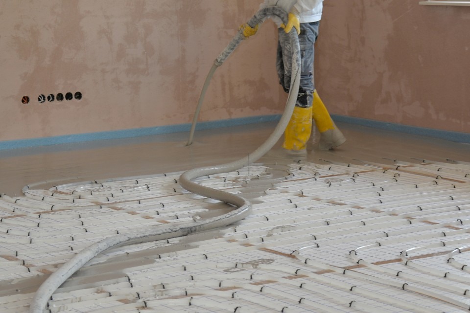 Wylewanie posadzki jastrychowej na ogrzewanie podłogowe, widoczne żółte gumowce pracownika i wąż doprowadzający mieszankę, ściany w surowym stanie.