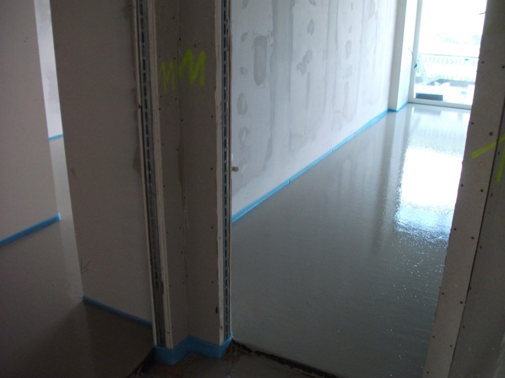 Świeżo wylana, gładka posadzka jastrychowa w trakcie schnięcia, zabezpieczona niebieską taśmą przy ścianach, widoczne metalowe profile przy ścianach.